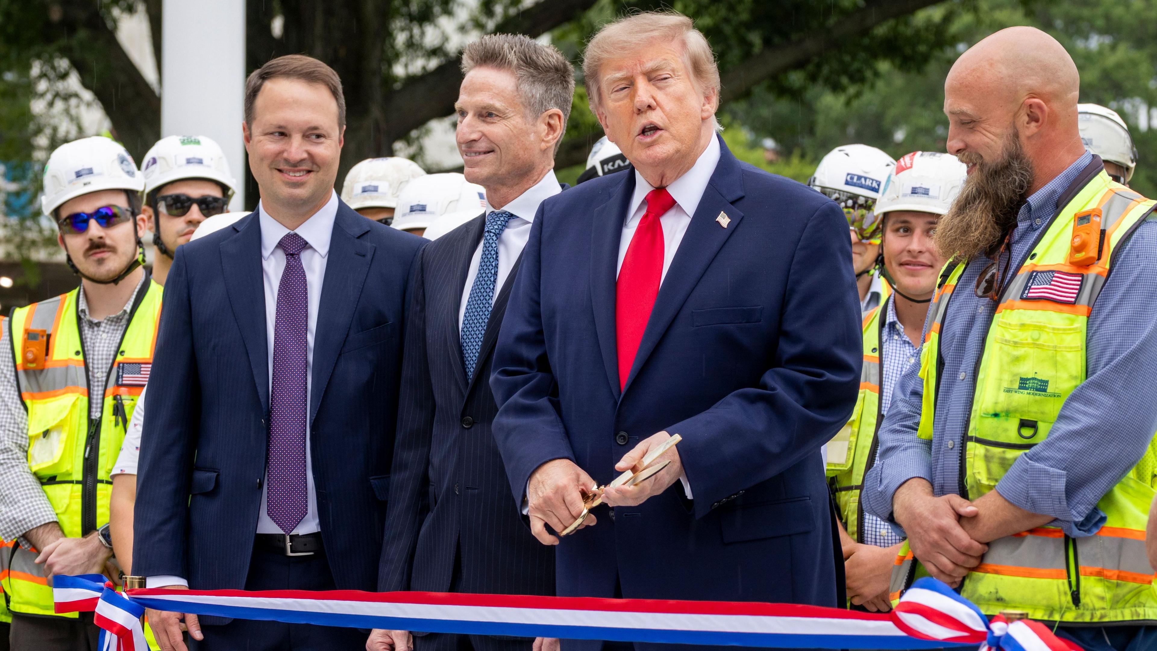 President Trump wears a navy suit and carries gold scissors to cut a ribbon in front of him