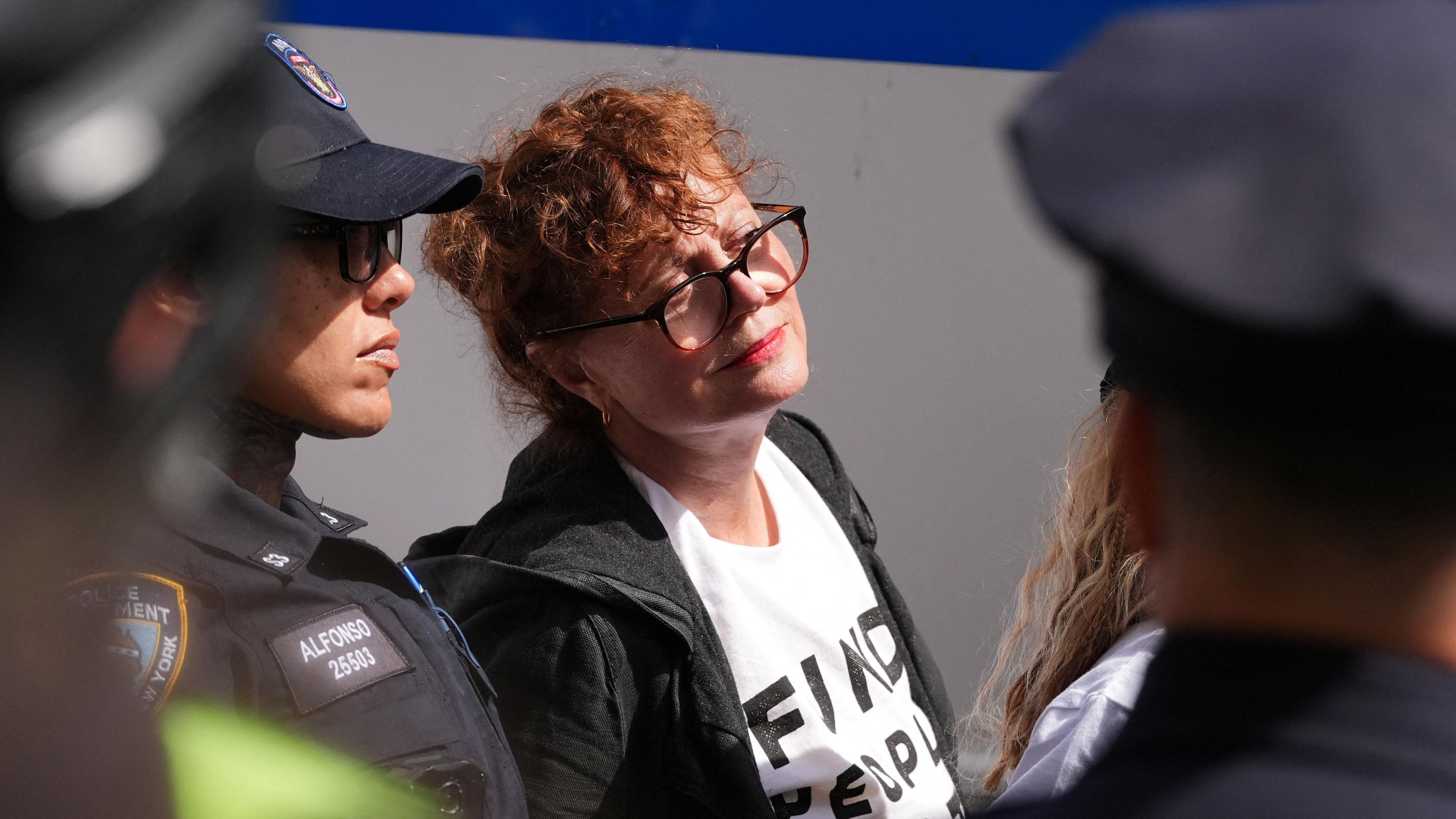 Susan Sarandon, whose ginger hair is tied up, wears glasses, a black hooded jacket and a white t-shirt, looks on as she is seen stood next to two police officers in New York City