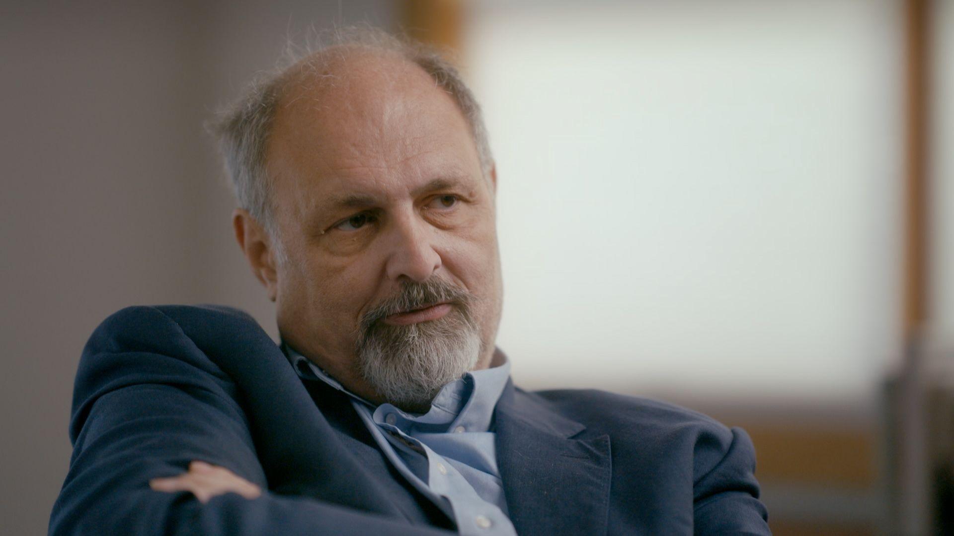 Jeffrey Breinholt, a man with greying hair at the sides of his head and a greying goatee beard, wearing a dark blue suit and a light blue shirt open at the neck, leaning back in his chair as he is interviewed