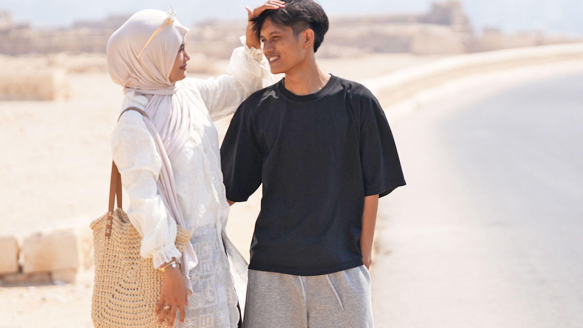 Zia has a straw bag over her shoulder, and wears a white blouse and pink hijab. She has her hand on Yusron's head. He has his arm around her and is wearing grey sweat pants and a black t-shirt.