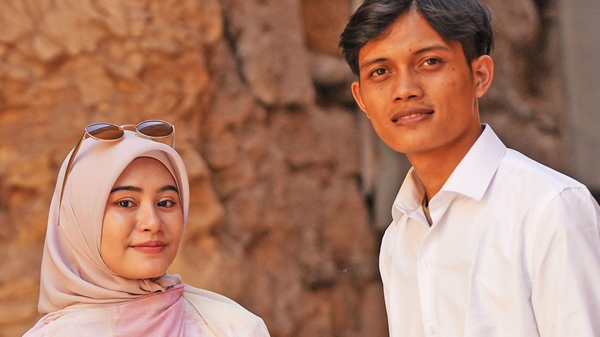 Zia and Yusron smile at the camera - Zia is wearing a dusky pink hijab and has sunglasses on her head, Yusron is wearing an open neck white shirt