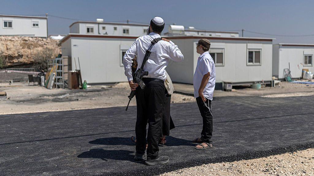 Three Israeli settlers stand outside houses after the reopening of a settlement at Ganim in the Israeli-occupied West Bank. One is a man armed with a rifle. Photo: August 2026