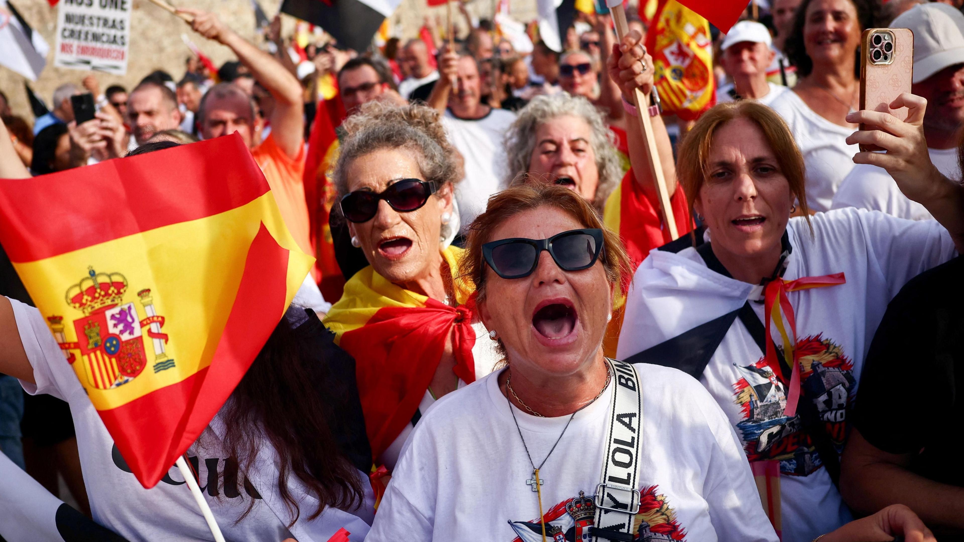 A woman at the front of a crowd chants with people behind and Spanish flags waving