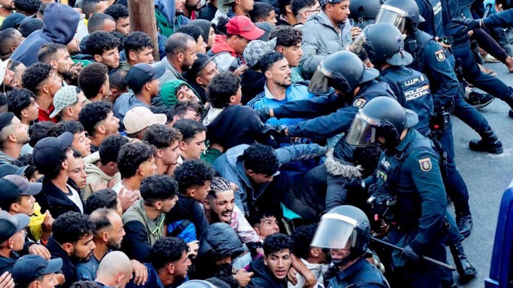 Police officers in riot gear push back a densely packed crowd of migrants during a chaotic transfer operation in Ceuta's port area