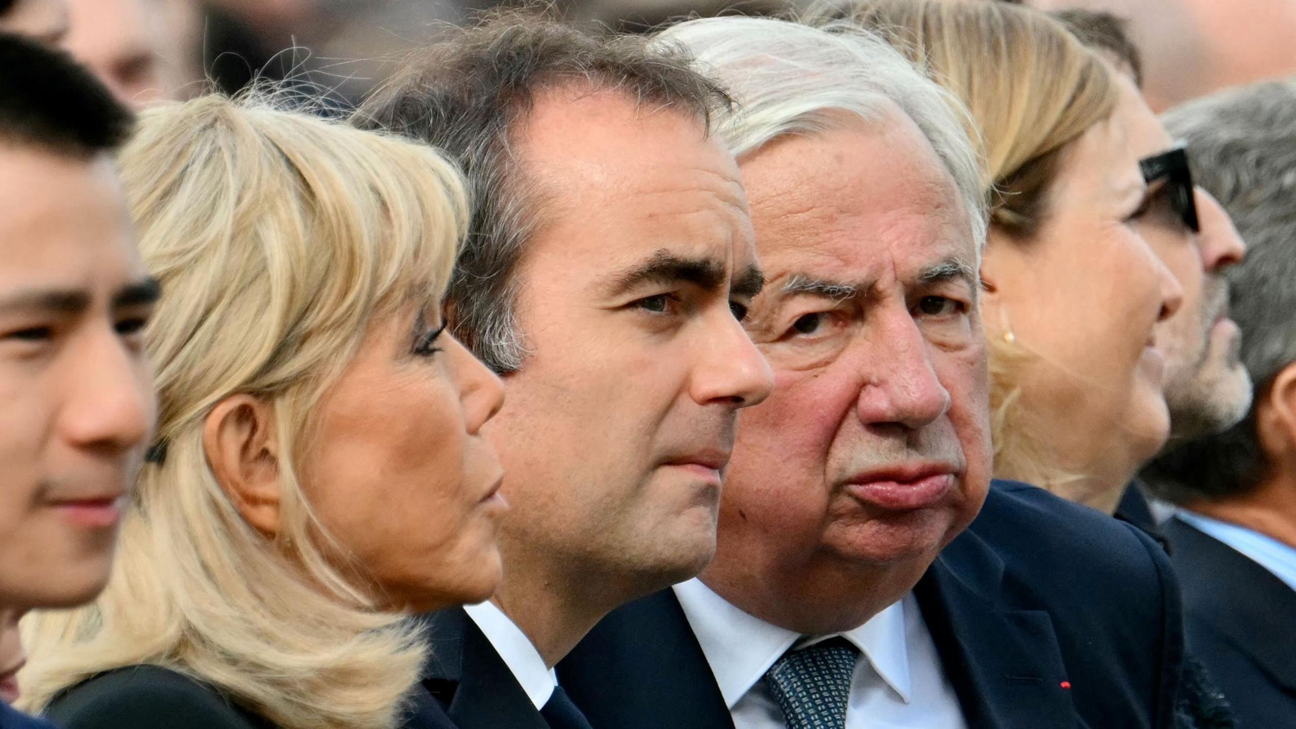 Brigitte Macron, Sebastien Lecornu and Gerard Larcher sit together and stare in different directions as they attend the outdoor mass in the centre of Paris.