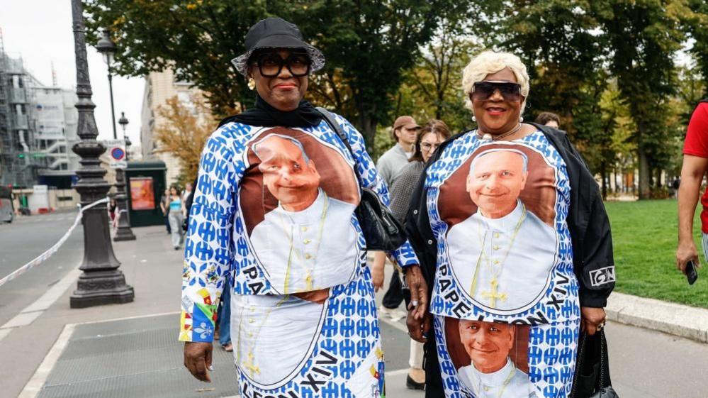 Two women wear outfits which have Pope Leo's profile on them alongside a blue pattern. Both of them have big smiles and are standing on a street in Central Paris.