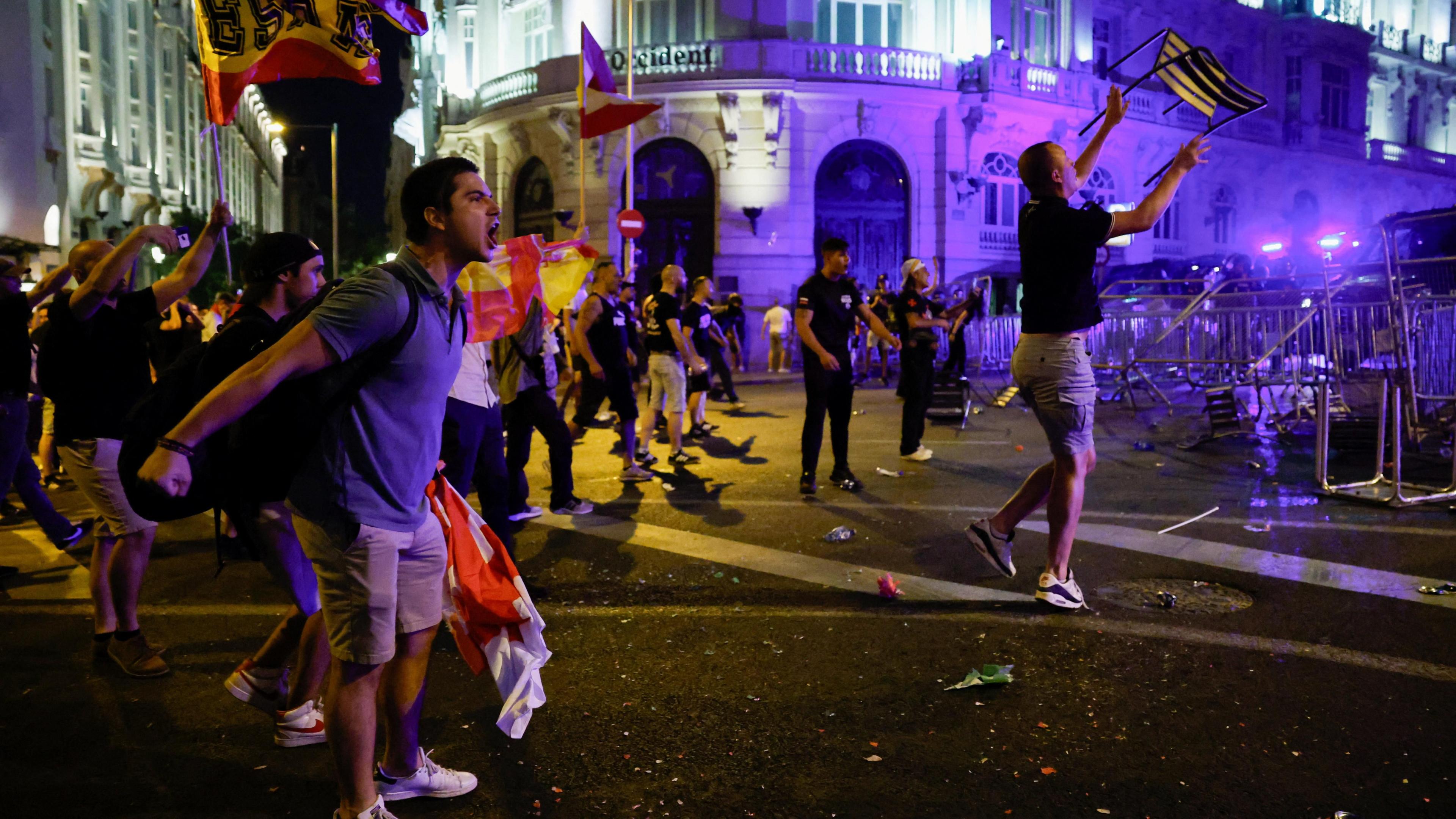 A man shouts at police with a barricade on the right and police lights visible. Other protesters wave flags.
