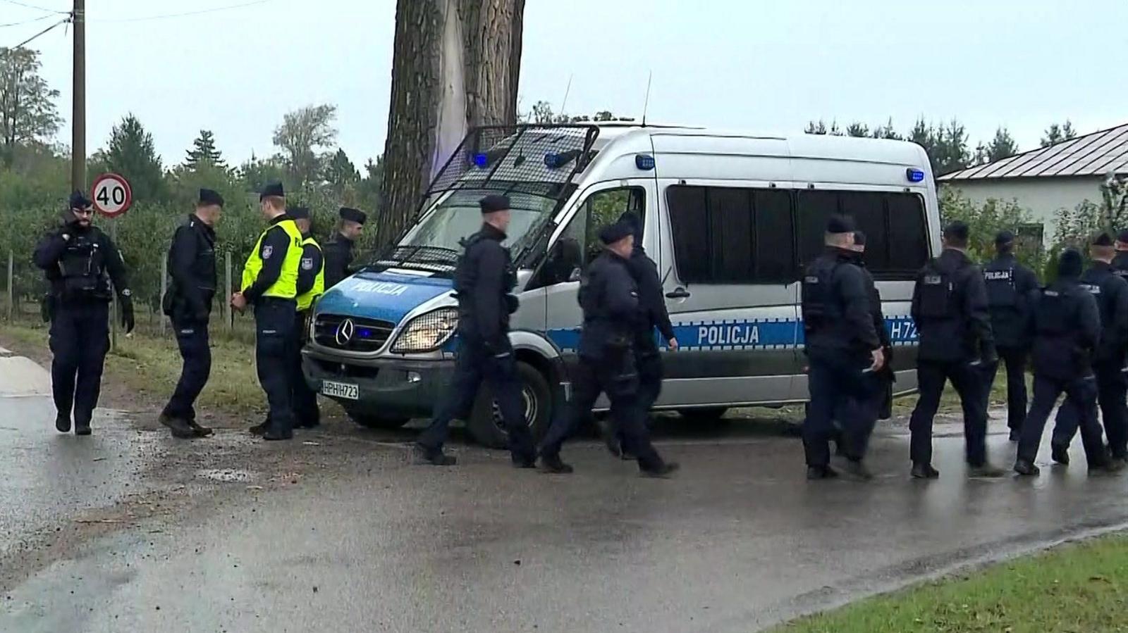 Police walk next to a silver police van near the telecommunications station in Wola Krobowska, Masovian Voivodeship province.