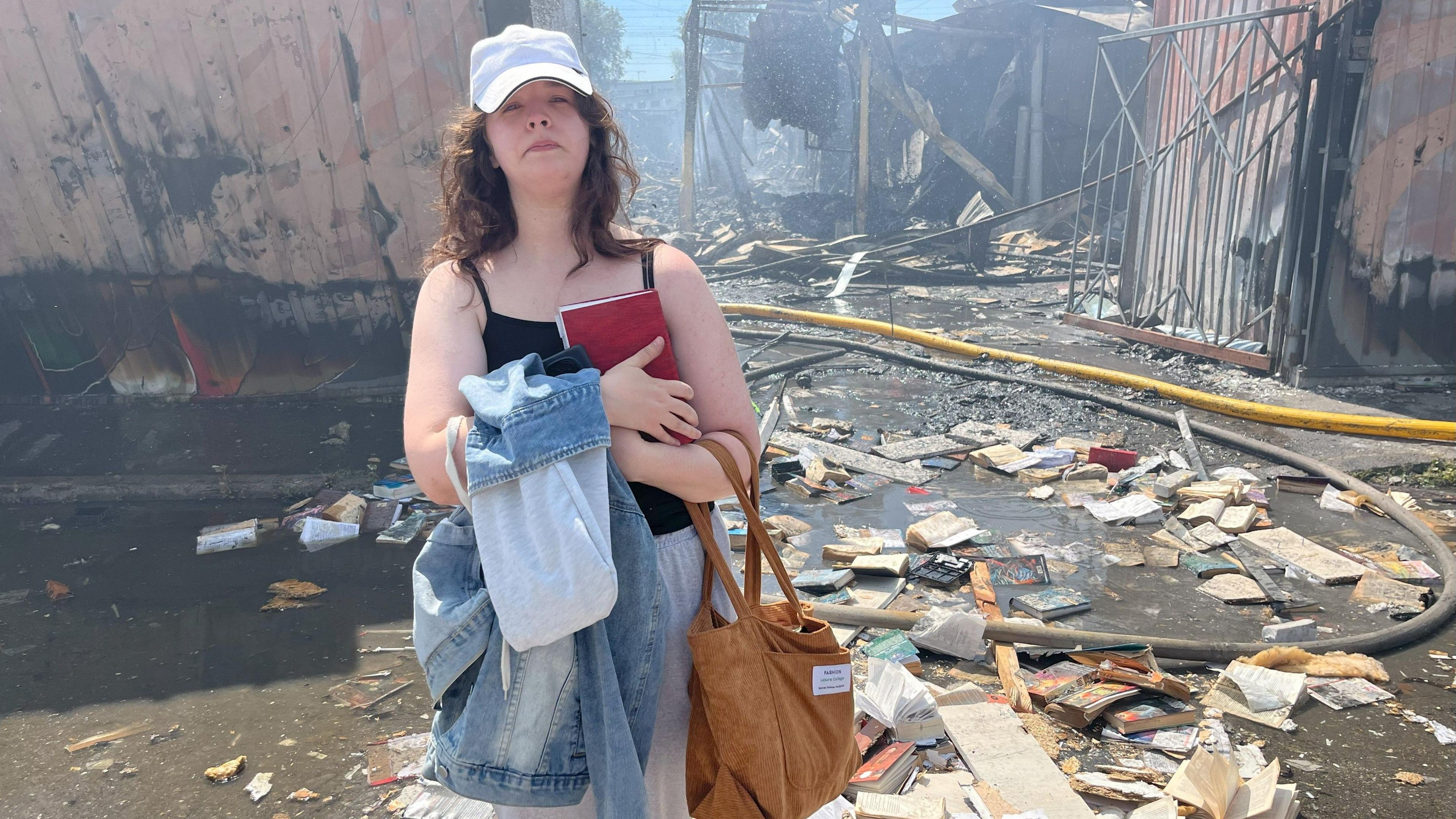A young woman clutches a book to her chest and looks sad. Behind her is a bombed-out market structure. Destroyed books litter the ground around her