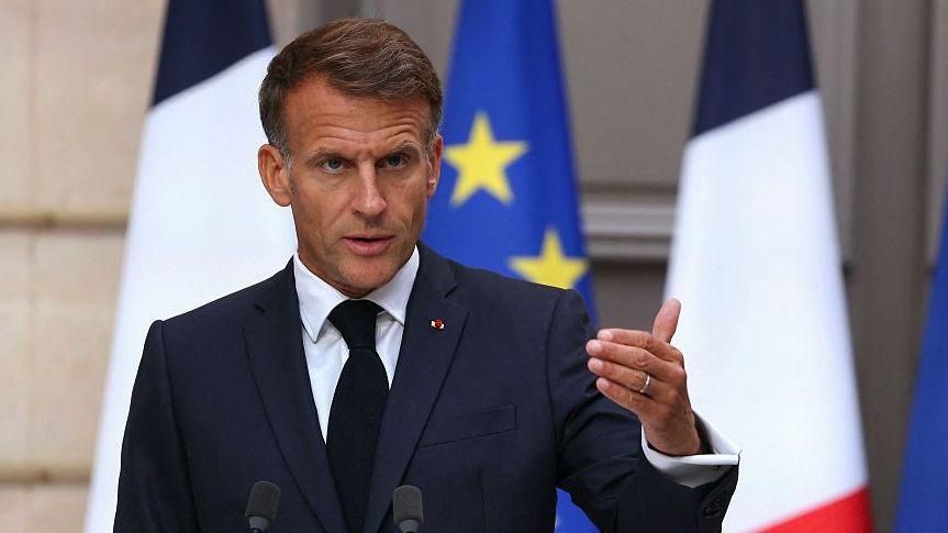 Emmanuel Macron, wearing a dark suit and tie, gestures with his left hand at a lecturn in the Elysee Palace with French and European Union flags behind him.