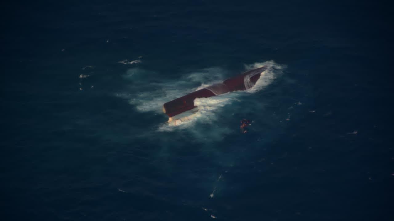 Aerial view of a capsized vessel partially submerged in dark blue waters with white foam surrounding the hull