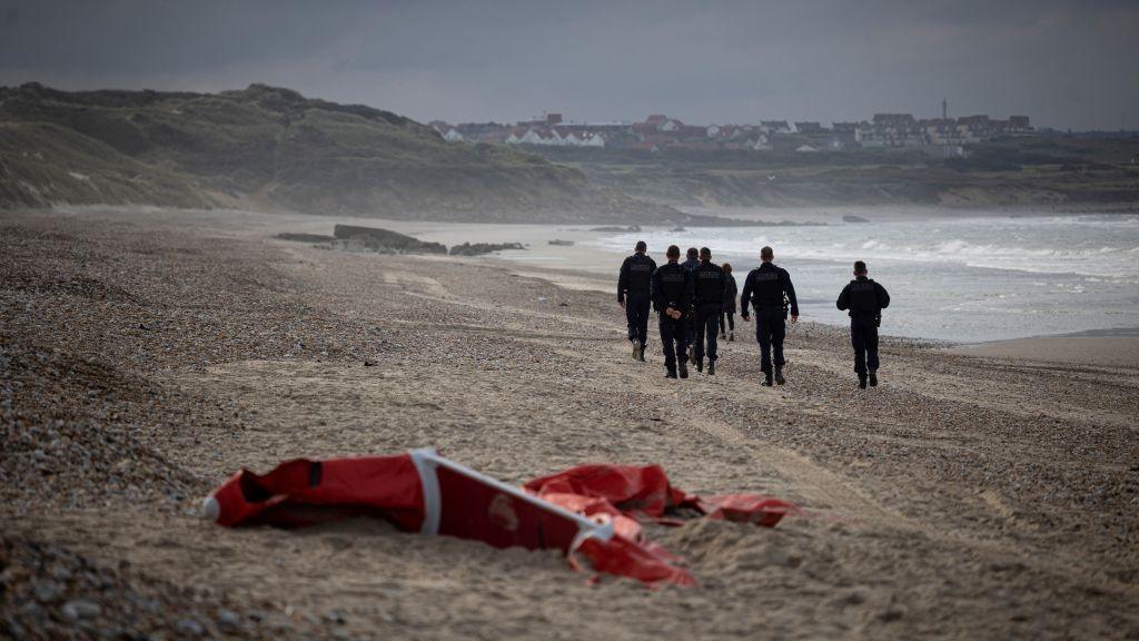 The remnants of an inflatable dinghy lie on a beach in northern France in October 2021. Police officers are in the distance patrolling on an overcast grey day.