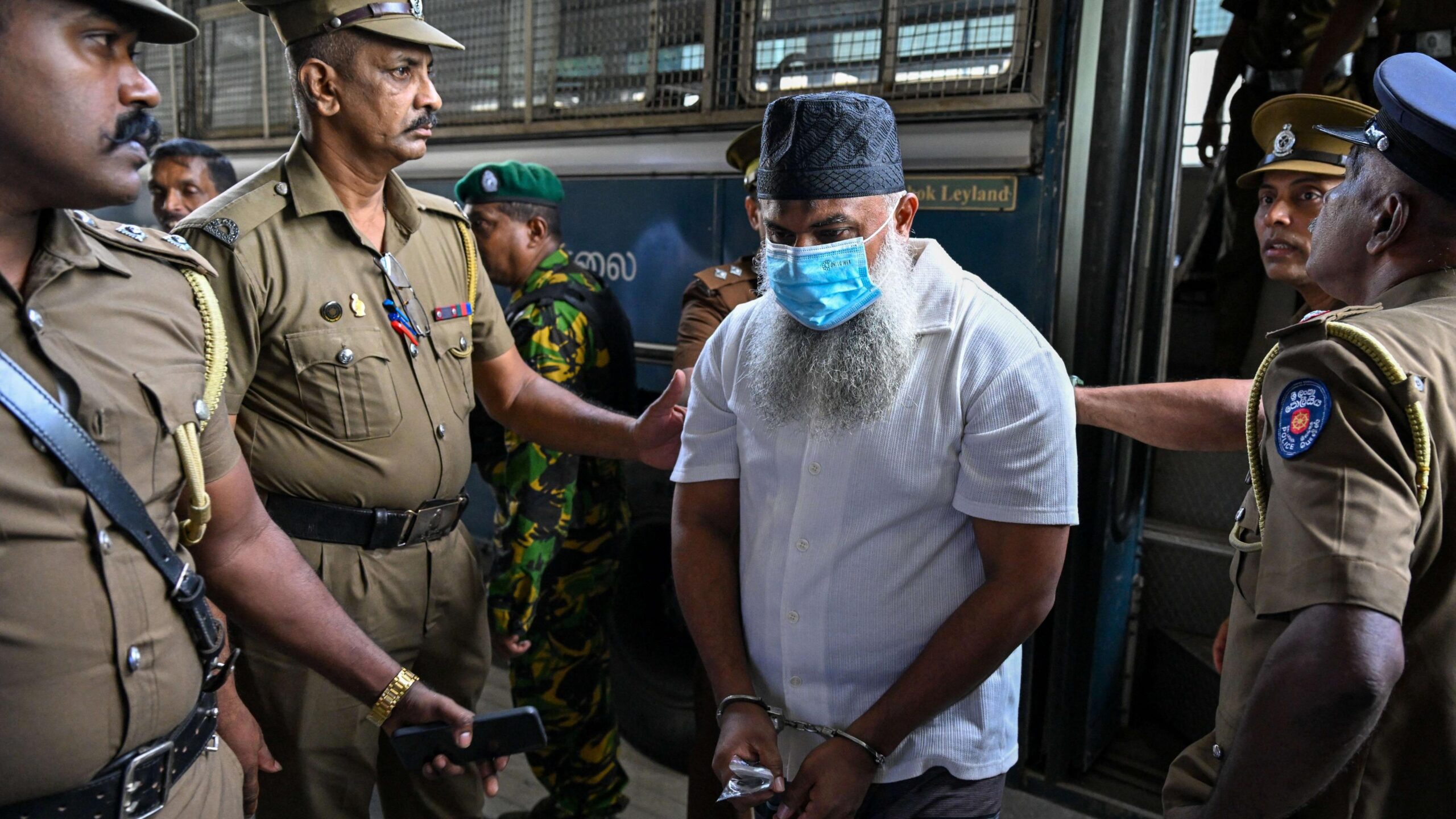 A man with a beard, skullcap and face mask being led out of a bus by four police officers