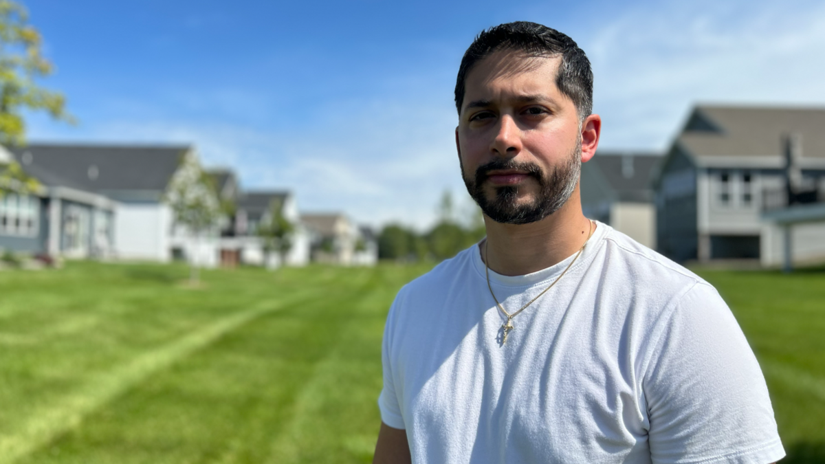 Gerald Bourguet, a white man standing in a field of a manicured lawn. He is wearing a white shirt and crucifix chain. There are homes in the background that are blurred.