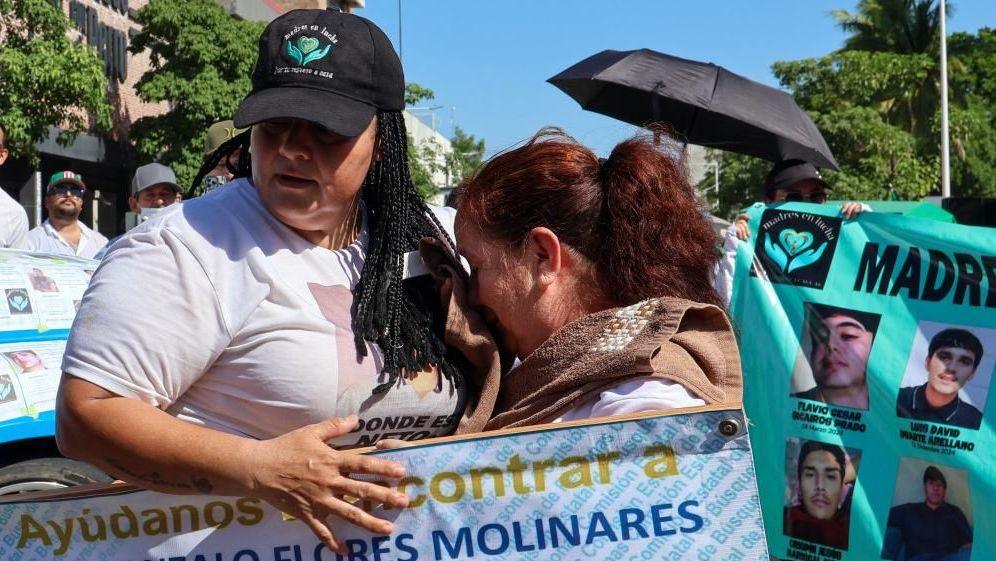 People take part in a march for peace in Culiacan, Mexico on 13 September 2026. A woman wearing a white T-shirt and a black baseball cap comforts another woman who is crying. The woman with the baseball cap is carrying a placard reading "help us find Jose Gonzalo Flores Molinares". In the background another protester is holding up another banner with the photos of missing people on it.