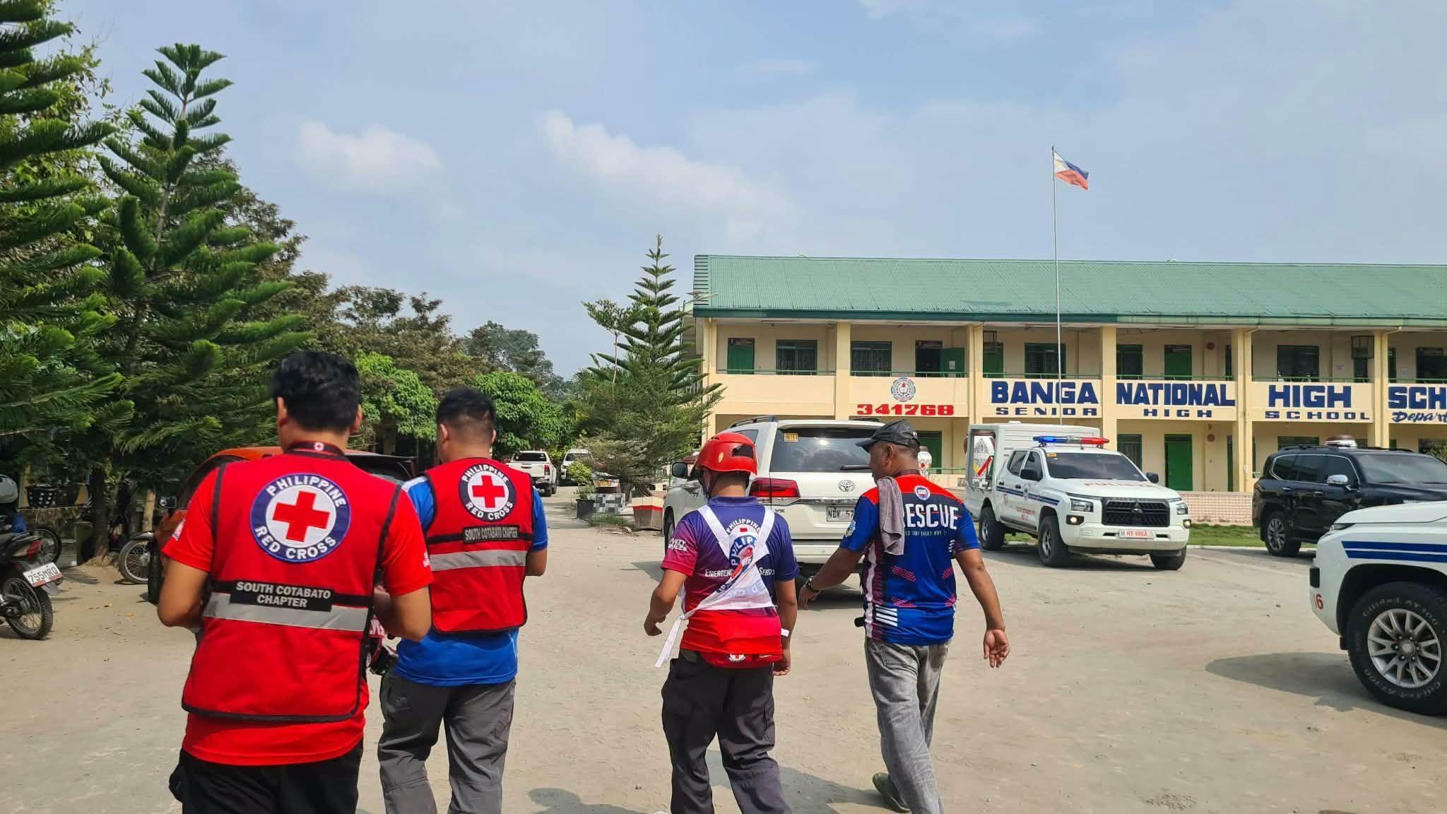 Members of the Philippine Red Cross and local rescue personnel walk across the grounds of Banga National High School in the Philippines. Several emergency and utility vehicles are parked in front of a two-storey school building displaying the words “BANGA NATIONAL HIGH SCHOOL.”