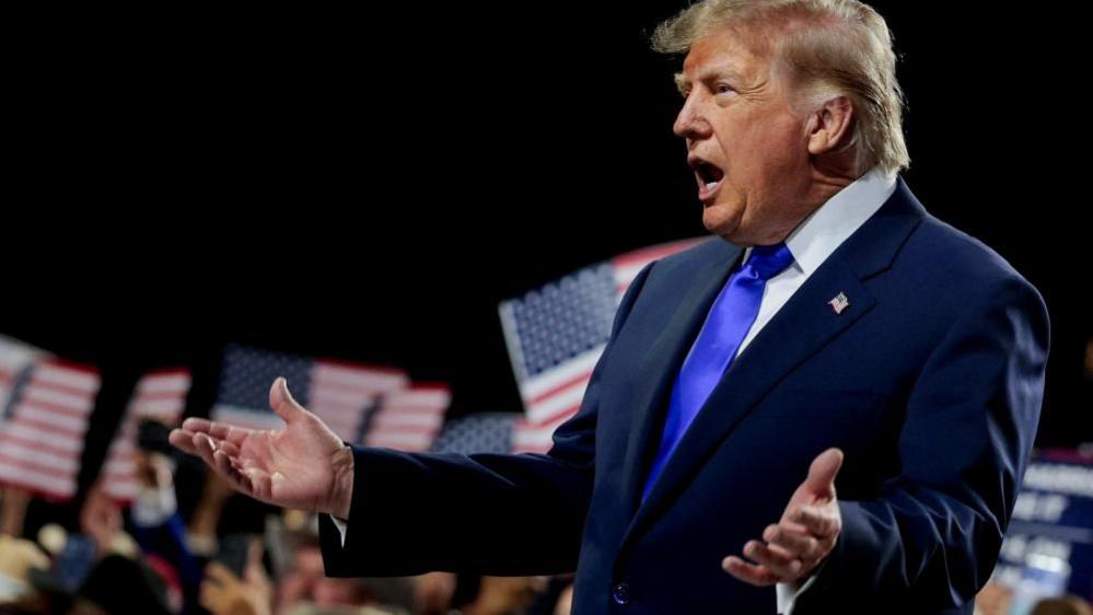 Trump wearing a blue suit and tie and white shirt opens his arms as he addresses the convention in Dallas