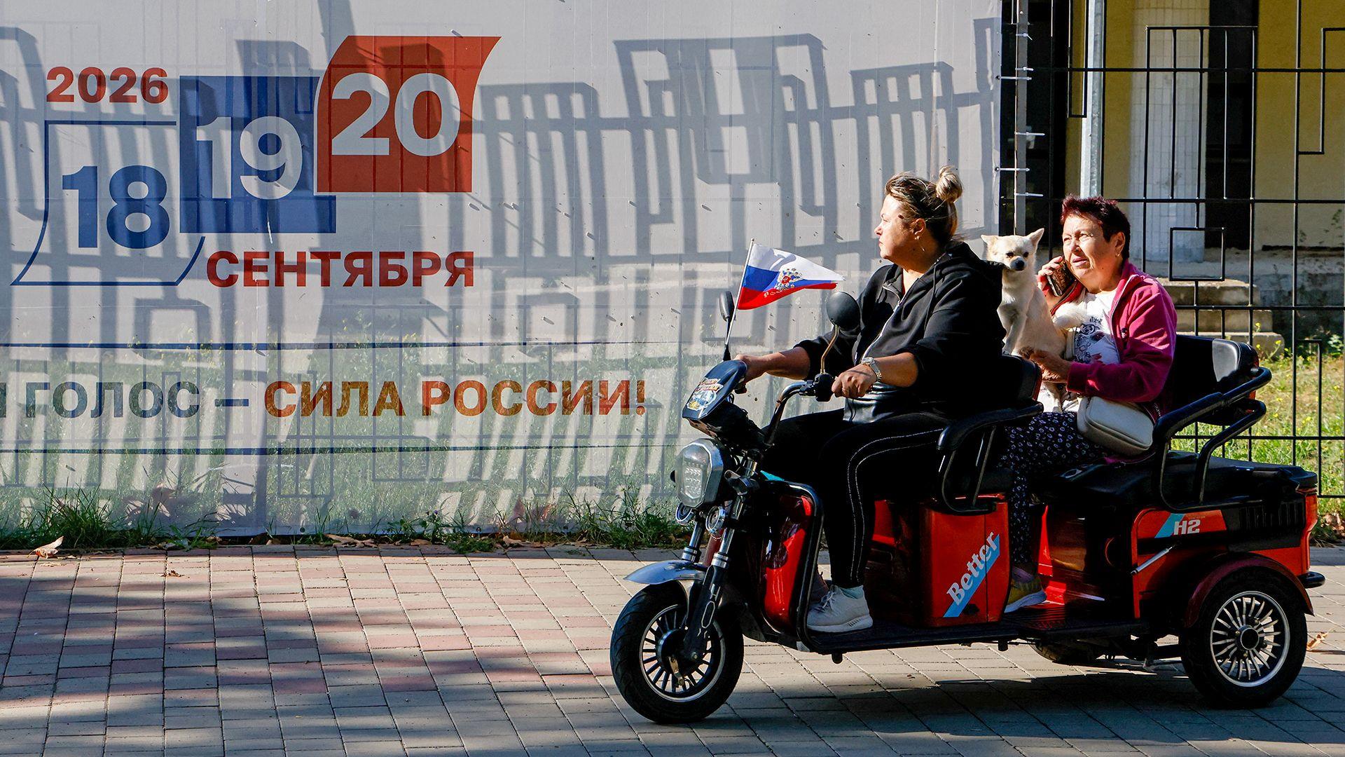 Two people ride an electric motorcycle along a street in front of a billboard advertising the upcoming elections, in Crimea on Tuesday.
