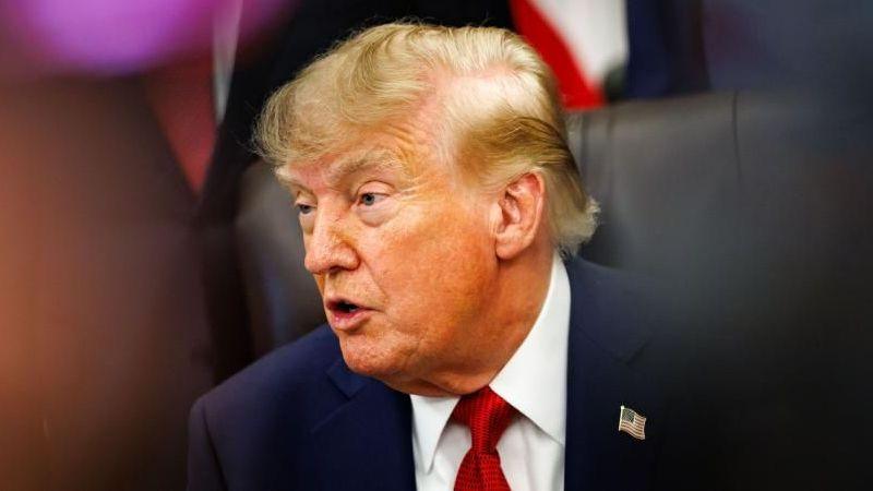 US President Donald Trump answers questions. He is wearing a dark blue suit jacket with a US flag on the right lapel, a white shirt and a red tie. He is sitting in a black high-backed leather chair. He is facing towards the left of the frame and is mid-speech.