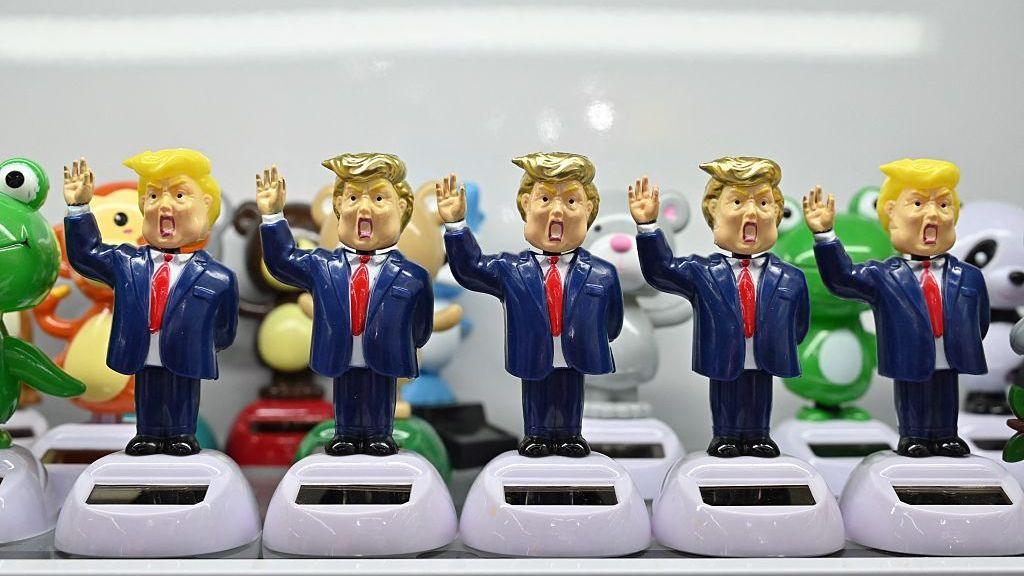 A row of five figurines styled as caricatured Donald Trumps stand on white bases with small solar panels. Each figurine wears a blue suit and red tie, with one arm raised in the air.