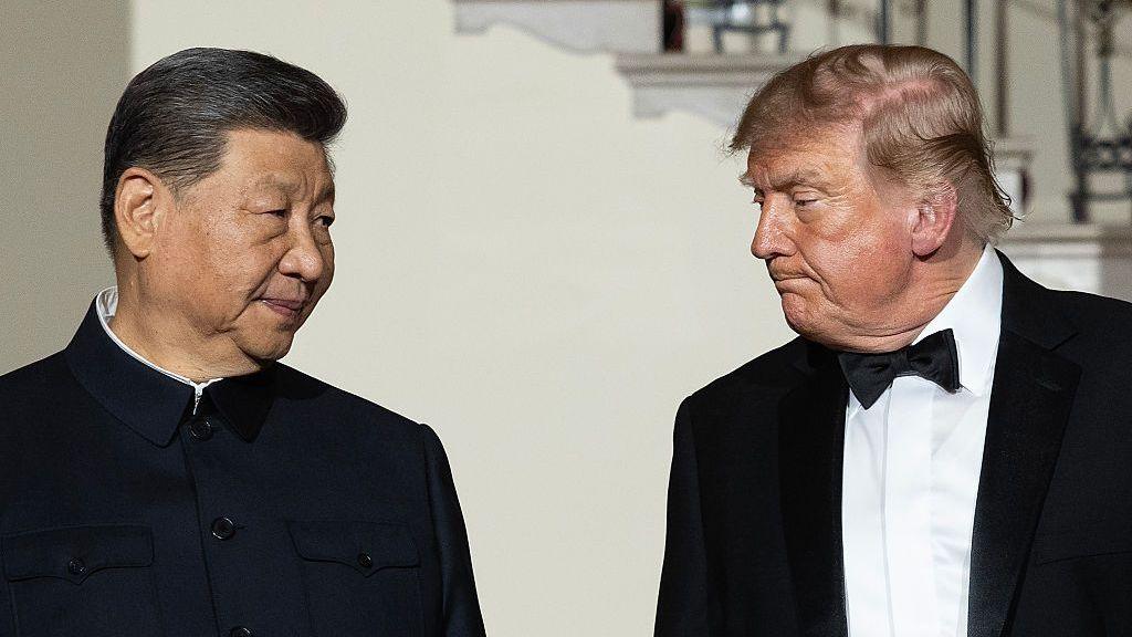 Xi Jinping and Donald Trump stand facing each other during a formal event, with Xi wearing a dark high-collared jacket and Trump wearing a black tuxedo with a bow tie.
