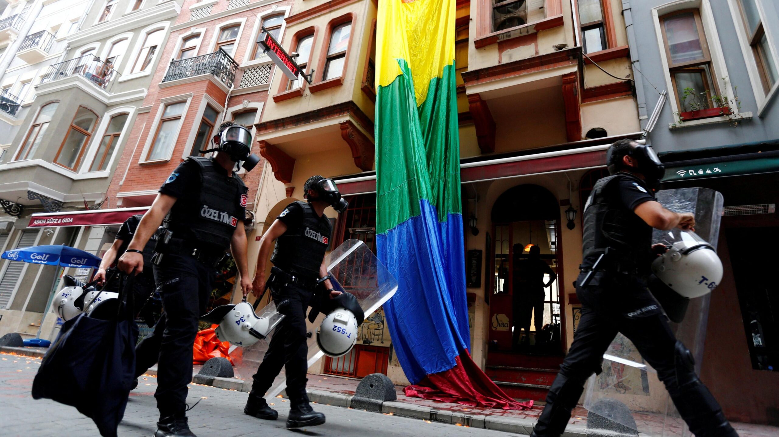 Riot police, with a rainbow flag in the background, chase LGBT activists as they try to gather for a pride parade, which was banned by the governorship in Istanbul. Officers are wearing black uniforms, wearing gas masks, and are carrying helmets and riot shields as they move through what appears to be a residential street.