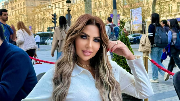 Sarah Khalifa wearing a white long-shirted with her folded left arm, the hand touching her long hair, sits by the side of a road inside a red-roped cordoned area, beyong which people walk on a road the other side of which has rows of sunbathed buildings