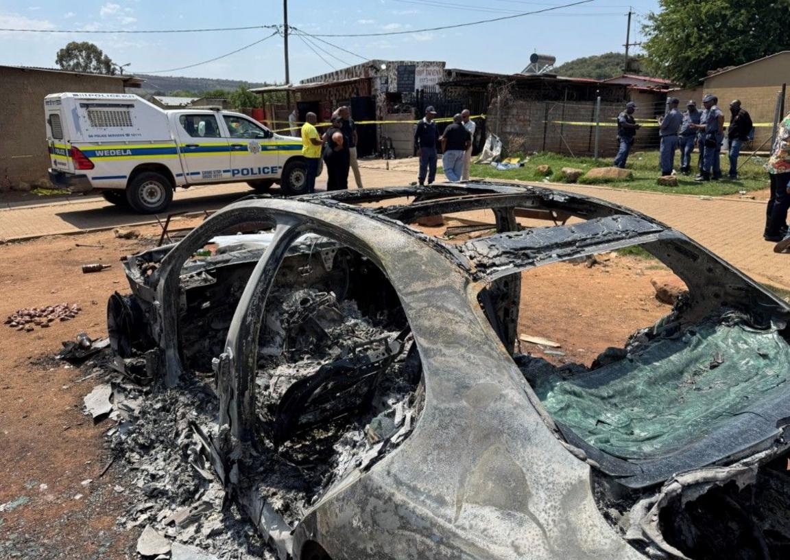 Shot of a burnt car in Johannesburg, while police officers and stand around it. A police van and a police cordon around a building can be seen in the background.