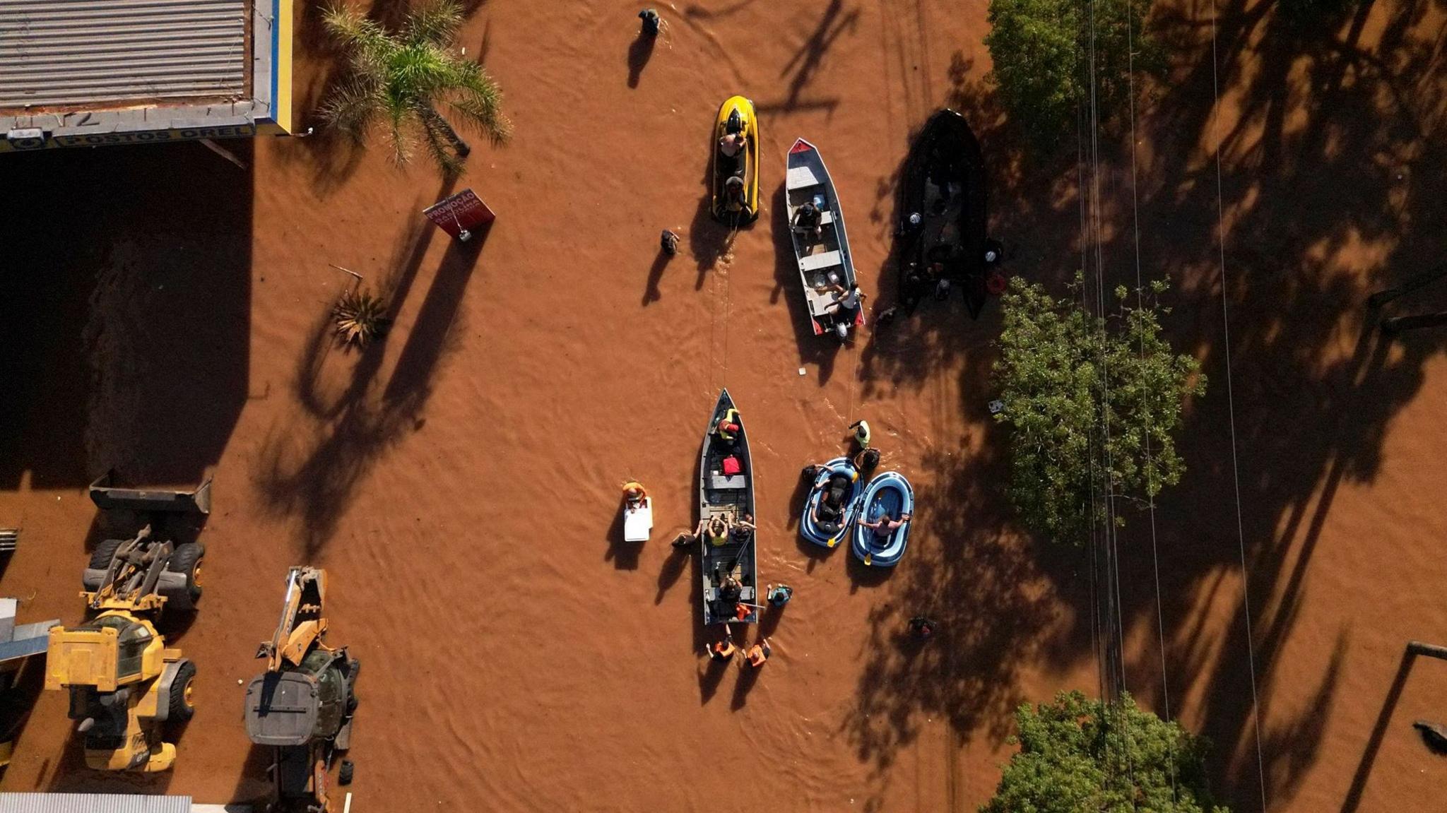 An aerial shot showing boats moving through brown floodwaters in a street. To the right are some trees and telephone wires, to the left is a corrugated iron building and some machinery.