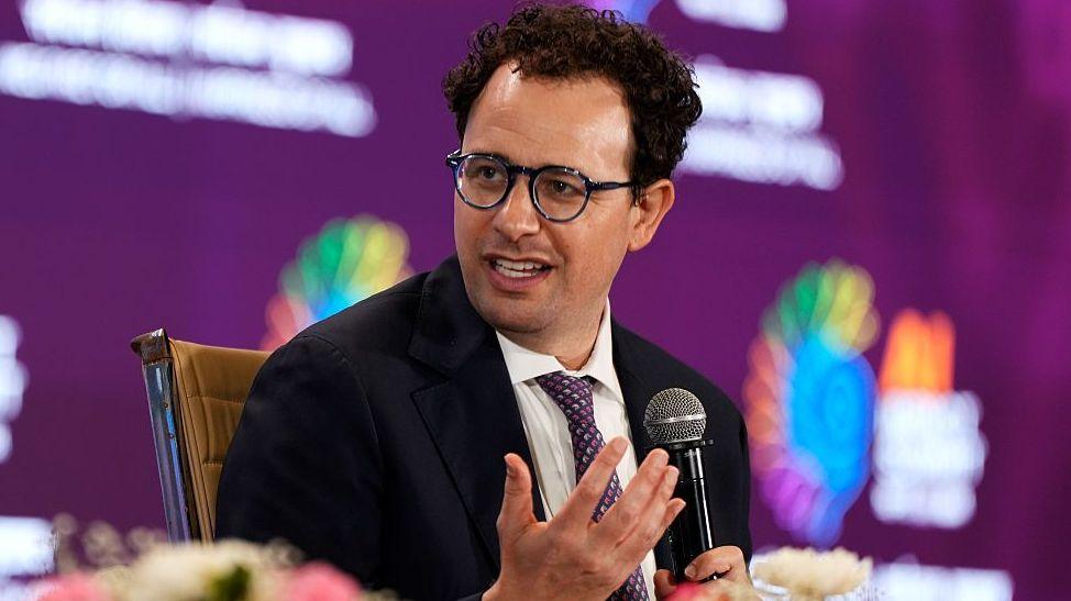 Anthropic boss Dario Amodei sitting and speaking at a conference, holding a microphone and wearing a suit with a purple tie