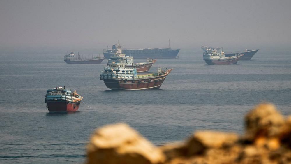 Several ships sit in a large body of water, with fog on the horizon and blurred rocks in the foreground.