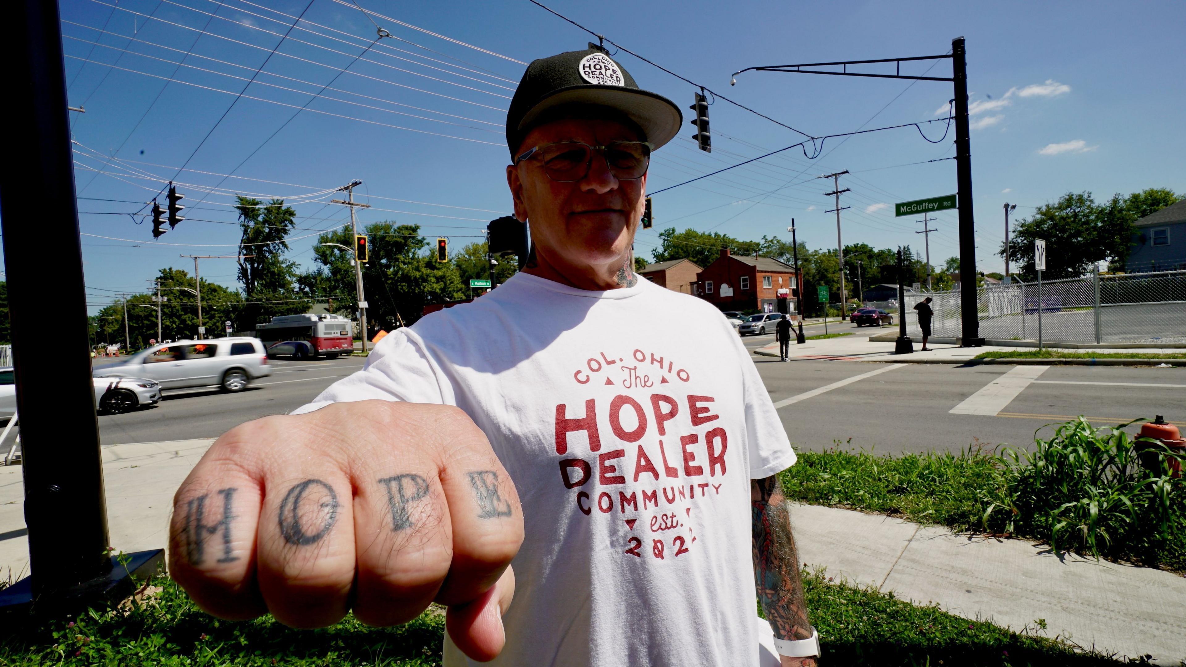 Scott Sanders, 65, wears a baseball cap and T-shirt that reads The Hope Dealer Community. He points his fist at the camera and on his knuckles reads Hope.