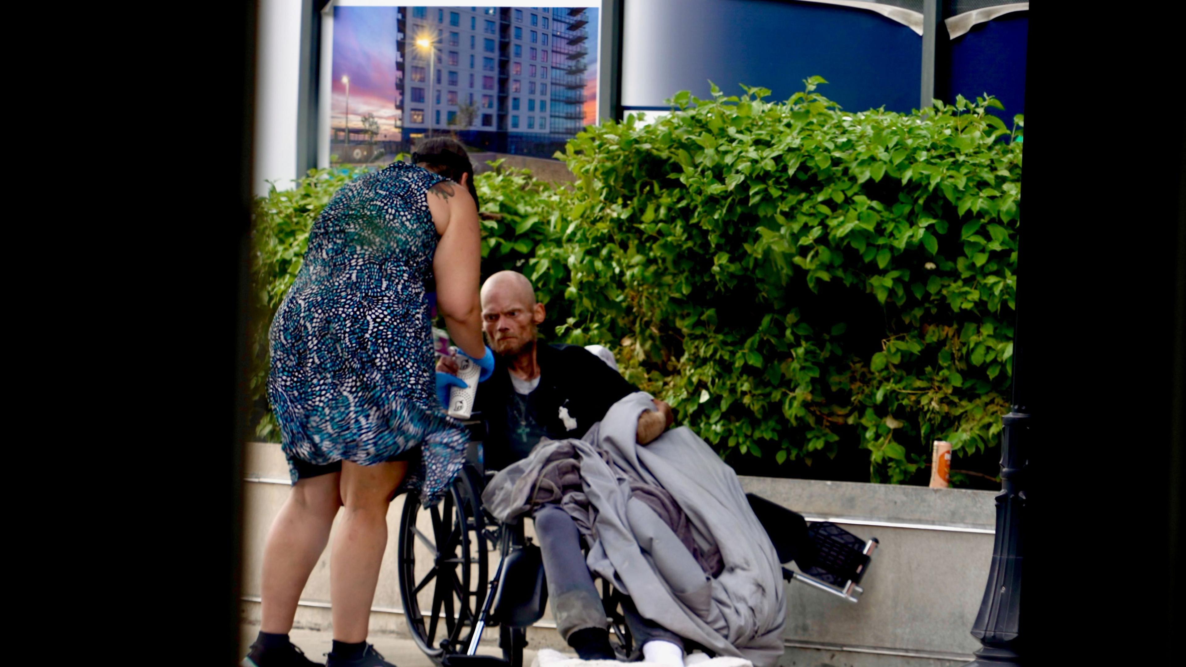 Karen, who wears a knee-length blue dress, tends to a gaunt-looking man in a wheelchair in an upscale neighbourhood