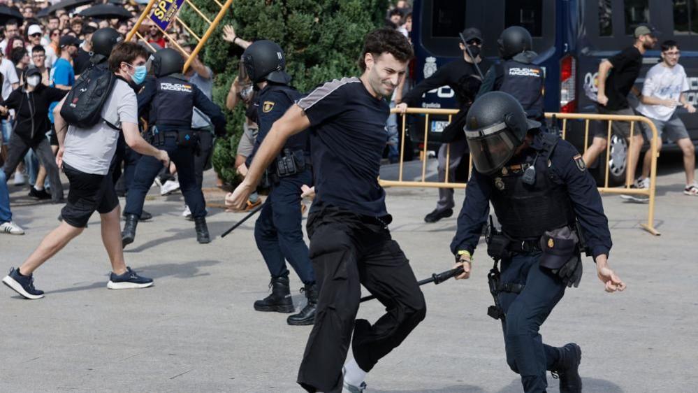 A riot police officer appears to be holding a baton near the back of a protester's legs. There are crowds of protesters in the background, and several riot officers in uniform with helmets. In the background, someone can be seen lifting part of a yellow fence.
