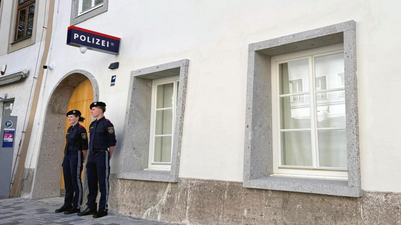 Two police officers stand outside a white-washed building with a police sign in German on the wall