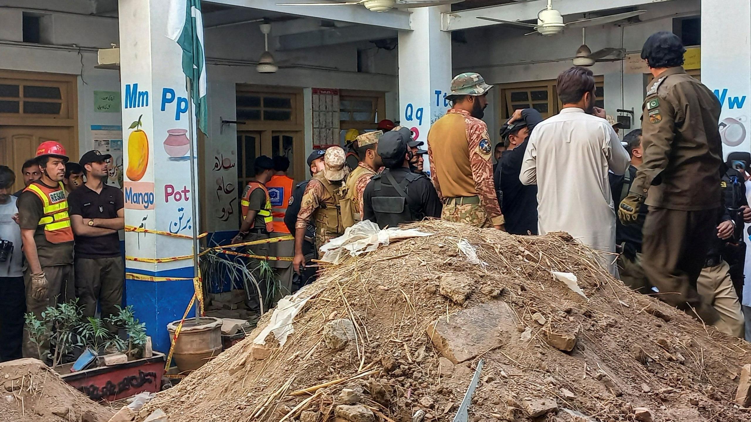Rescue workers, police and security personnel gather inside a school building around a cordoned-off area beside a large mound of excavated soil. Educational murals and a Pakistani flag are visible on the pillars