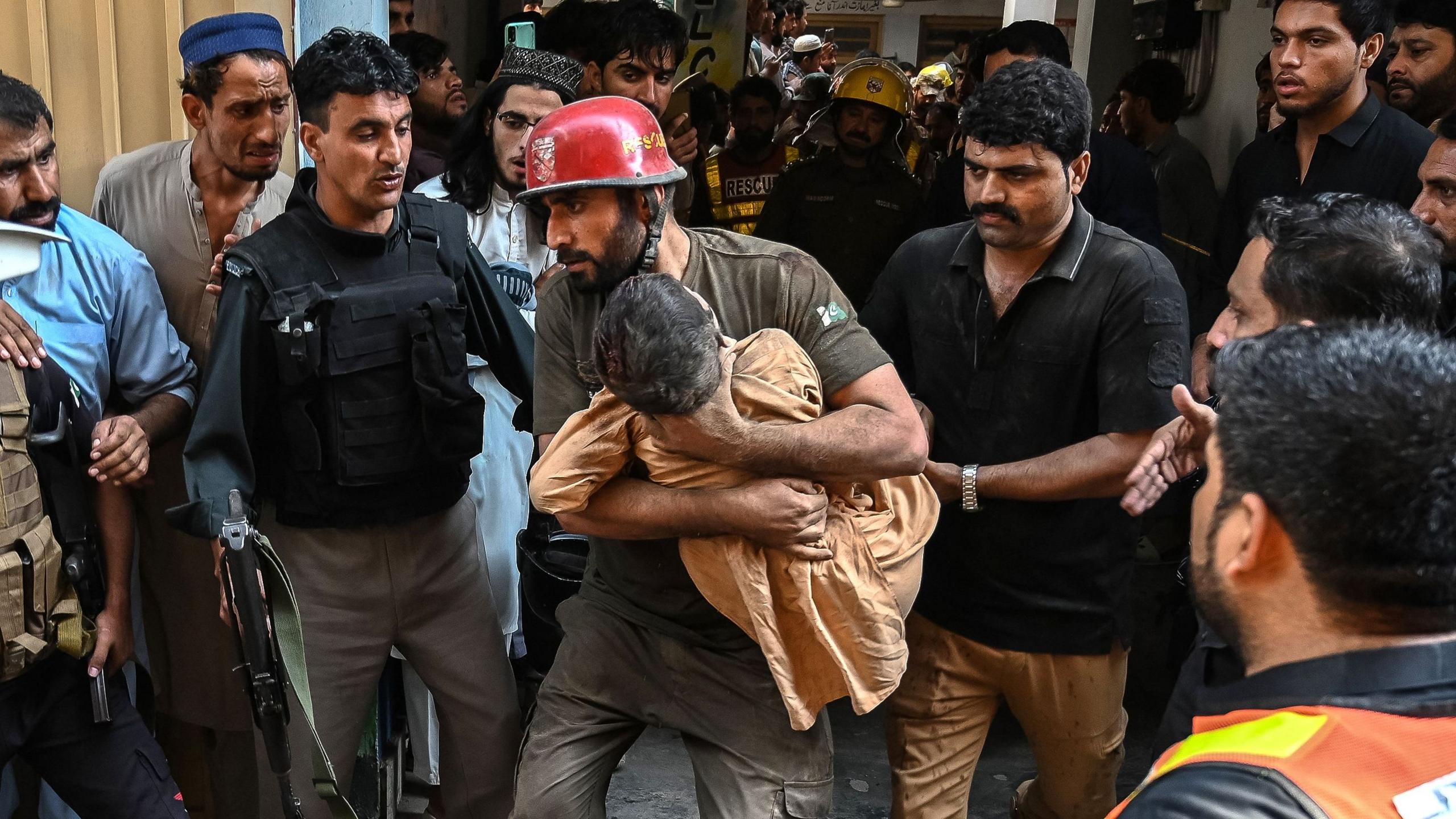 A rescue worker wearing a red helmet carries a small child as police officers, emergency responders and onlookers surround them.