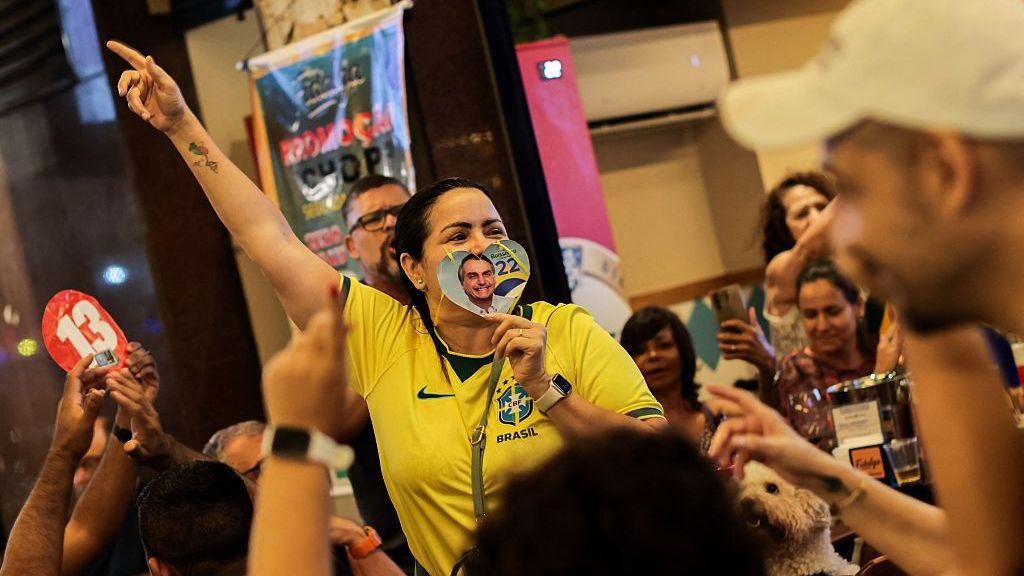 People gather around a woman in a yellow Brasil football jersey, raising one hand in the air, as she holds over her mouth a love heart-shaped cutout showing Jair Bolsonaro's face next to the number 22