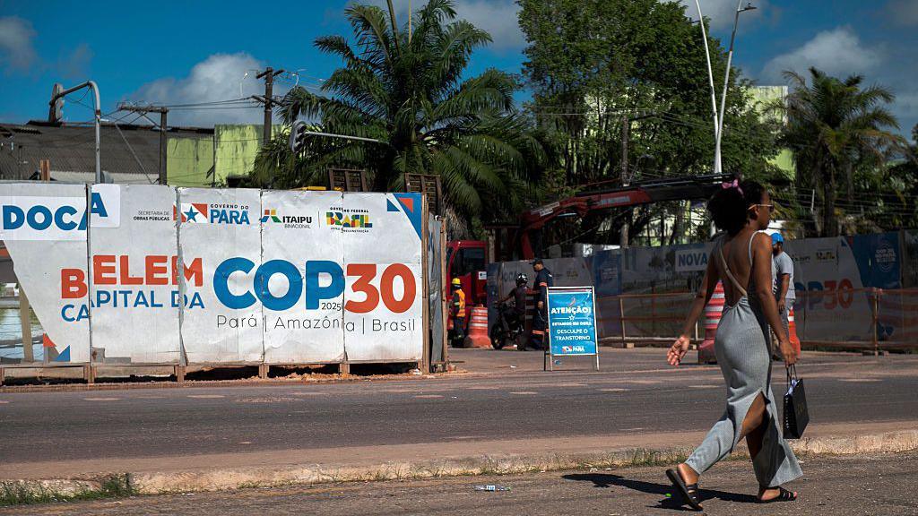 A woman walks past an infrastructure project under way for COP30 in Belem. She has her hair up and is wearing a grey dress and carrying a black bag. Misaligned posters in white, blue and orange read "Belem Capital Da COP30".