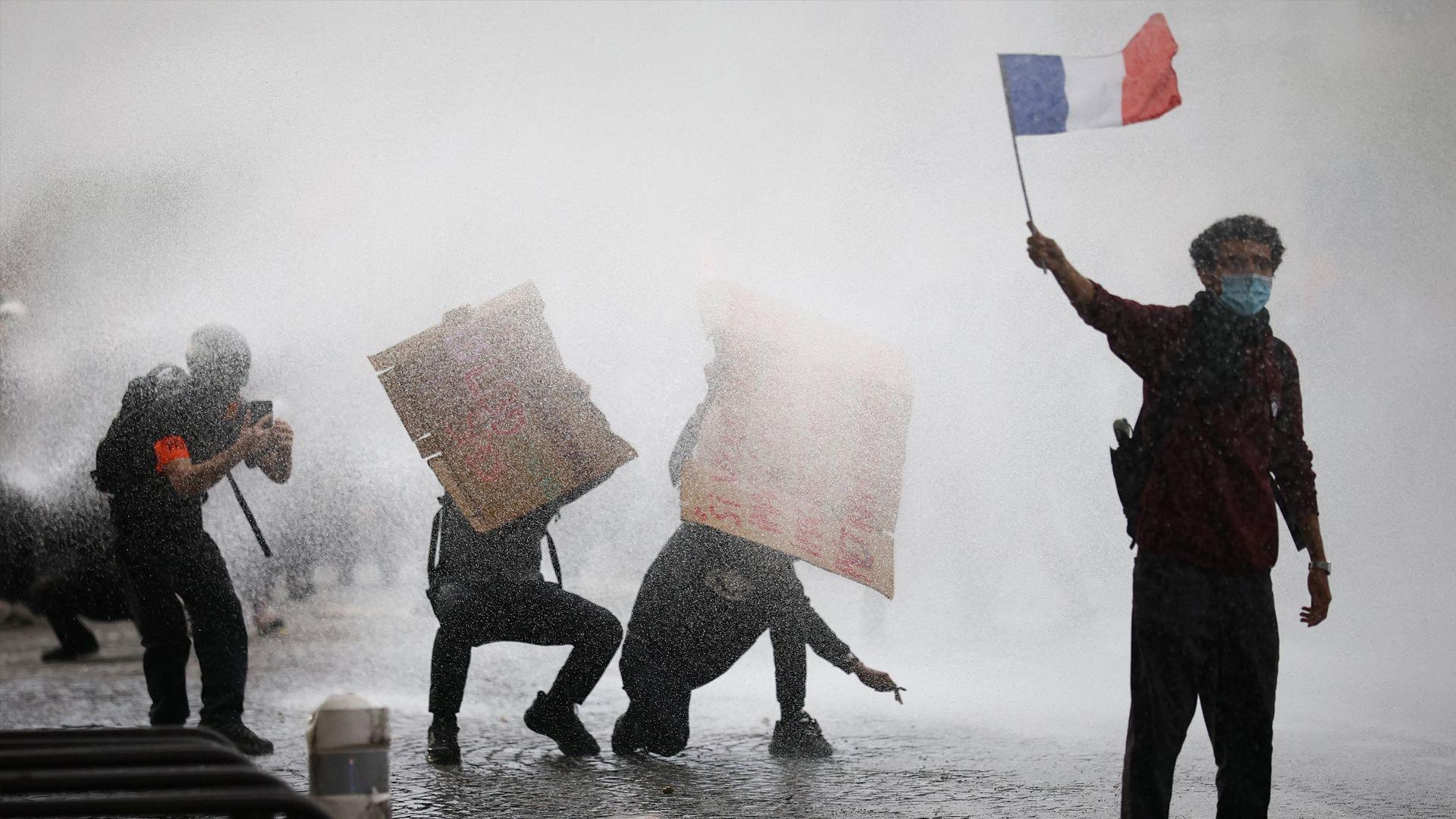 A masked protester holds a French flag as French police use a water canon amid clashes during a demonstration by French high school and university students in Paris.