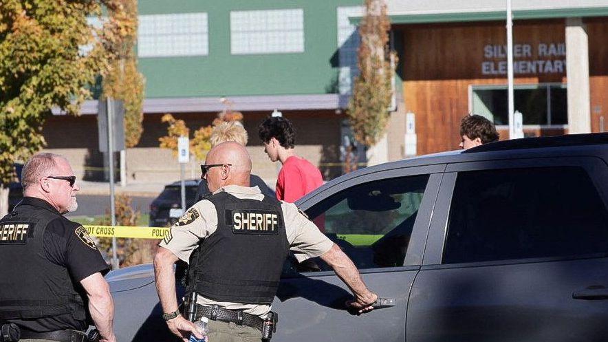 Two men stand in the lower left corner wearing bullet-proof vests that say "sheriff" on them. In the background is a green building with wooden panelling, with the name of the school on it.