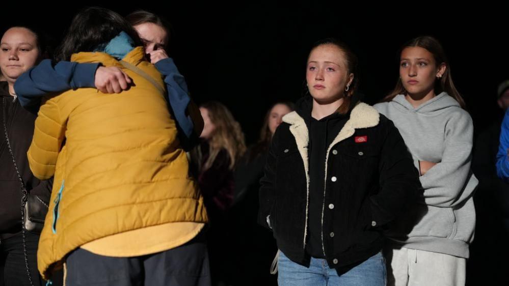 Two people hug each other while three women look on. All of them look sombre and are wearing light jackets.