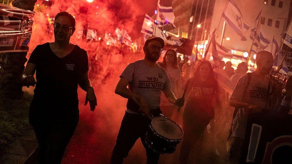 Protesters bang a drum and light red flares. It is night.