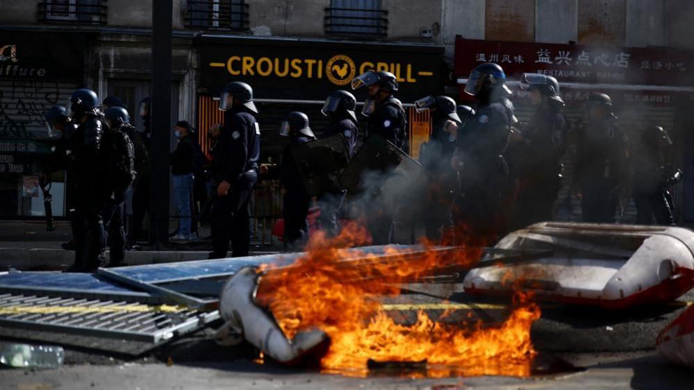 Riot police stand near burning street barricades during student protest unrest in France, as flames and smoke rise from debris blocking a city road