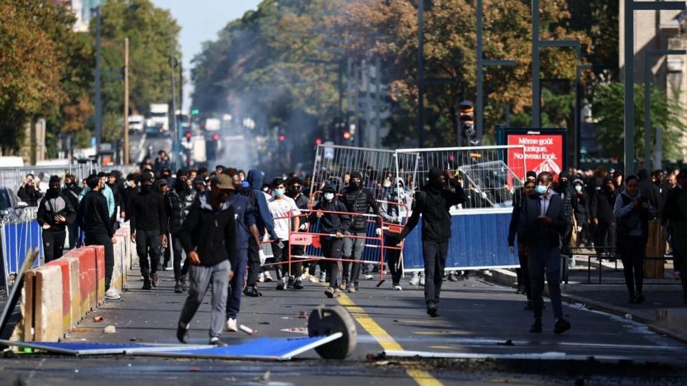 Students march along a street lined with barriers during protests in France, with debris on the road and smoke visible in the background