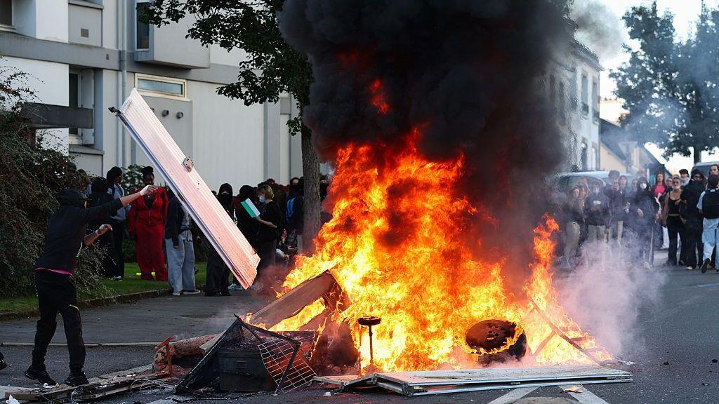 An enormous fire is seen outside a school as protesters look on from behind
