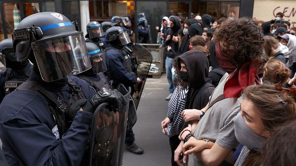 A close-up shot of a row of police in riot gear on the left of the frame, facing a row of young protesters on the right. The police officers wear helmets with visors, mouth coverings, vests and shields. Their uniforms are black and dark blue. The protesters wear street clothing like hoodies and t-shirts and have their mouths covered with scarves and masks.