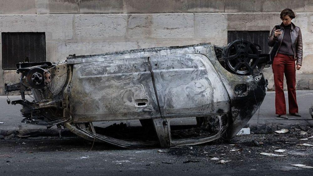 A woman takes a photo of the wreckage of a burnt-out car in central Paris
