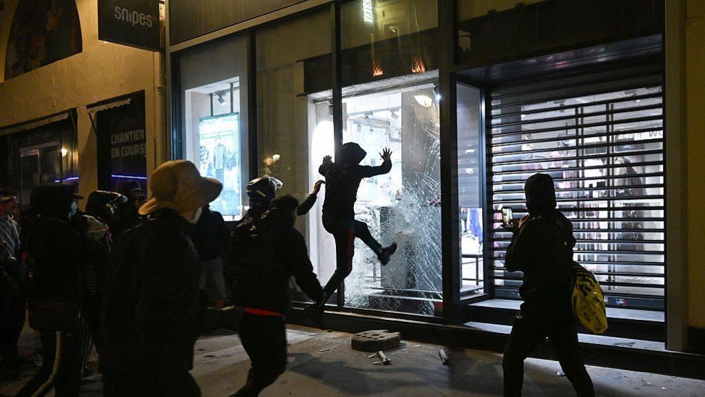 People try to destroy the window of a Snipes store near Chatelet, Les Halles in Paris as riots broke out. There are a group of onlookers looking at someone smashing a shop window. They are all wearing dark clothing and have their faces covered. One person is filming it on a phone.