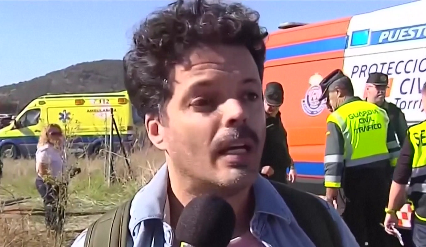 A man with black hair and moustache in front of a micophone, with emergency vehicles and personnel wearing yellow vests in the background
